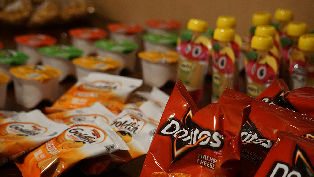 Close-up of assorted snack items on a table, including individual bags of Doritos and Goldfish crackers, fruit pouches with yellow caps, and small yogurt cups in assorted flavors.