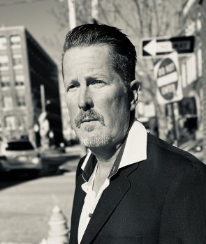 Jeffrey Thomson
 stands outdoors on a city street, wearing a dark blazer over a light collared shirt. The black-and-white photo shows him looking off to the side, with buildings and street signs softly blurred in the background.