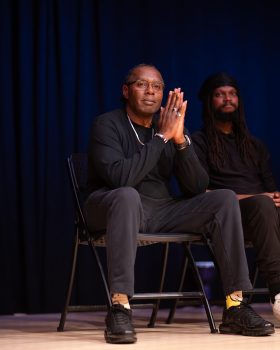 Joseph Jackson sits on a stage chair with his hands clasped in a thoughtful pose, wearing a black shirt and dark pants. A blue backdrop fills the background, with another seated person partially visible behind him.
