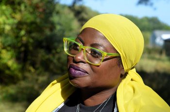 Samaa Abdurraqib looks slightly upward outdoors, wearing a bright yellow headscarf and translucent green glasses. Sunlight highlights her face, with trees and a building softly blurred in the background.