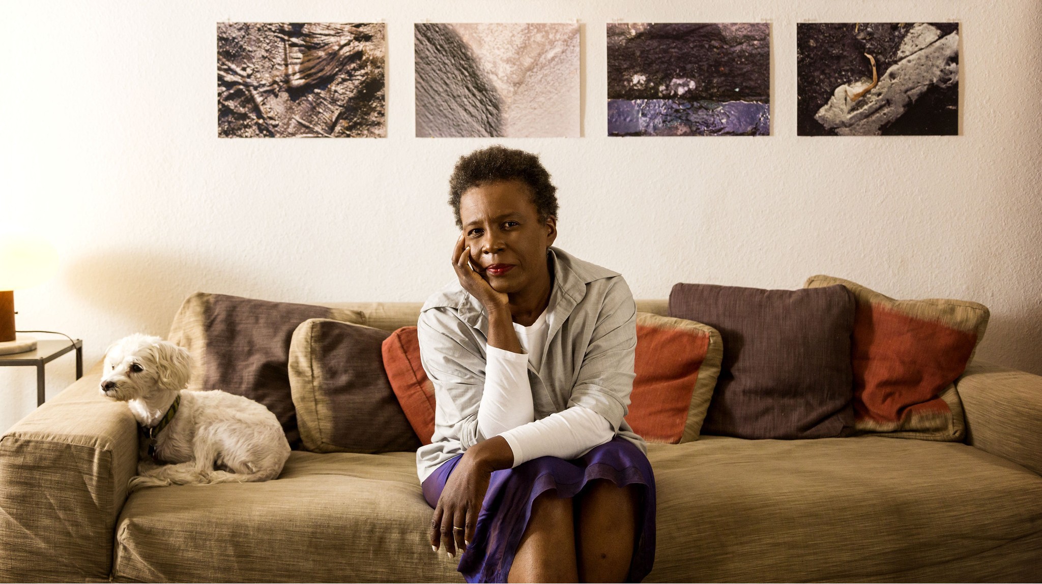 Author Claudia Rankine sits on a couch, elbow on knee and chin resting on hand, her miniature poodle rests its head on the arm of the couch.