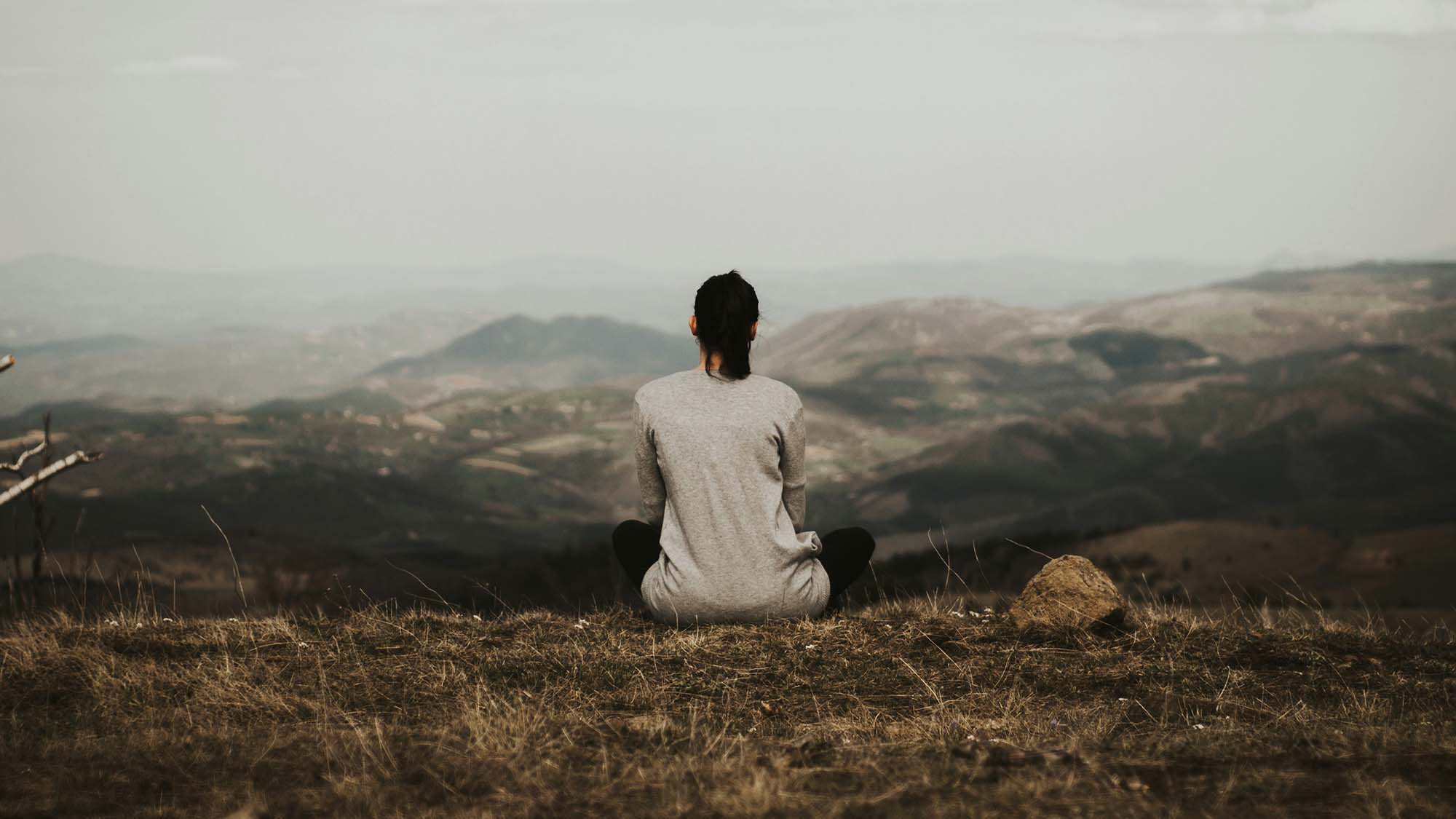 Person sitting cross-legged on a hillside overlooking a wide mountain landscape.