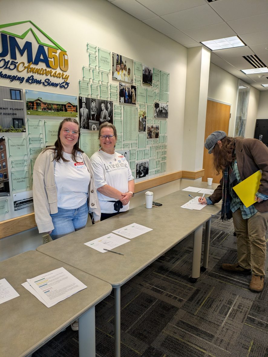 Two volunteers welcome participants at a registration table while an attendee signs in.