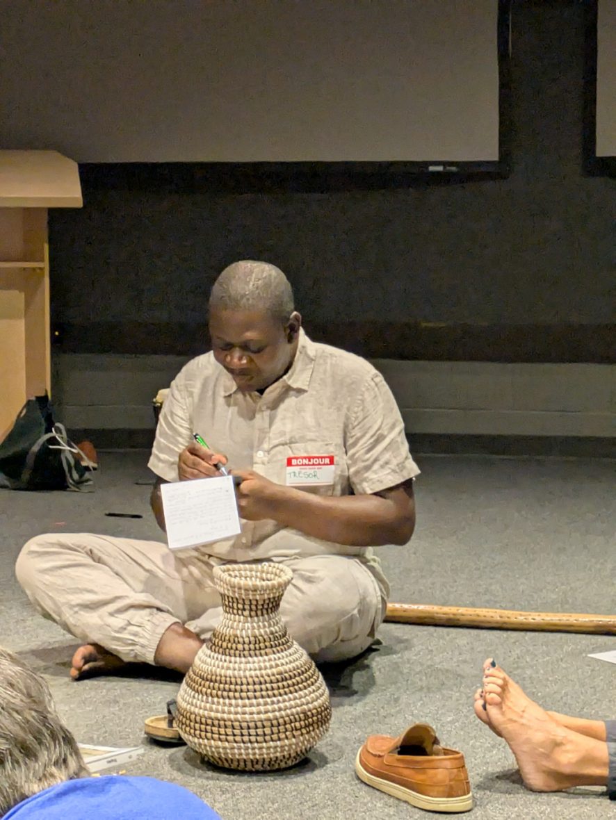 Participant seated on the floor during a cultural activity, writing on a card beside a woven basket.