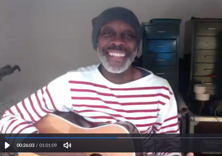 A smiling man wearing headphones and a red-and-white striped shirt holds an acoustic guitar during an online video presentation.