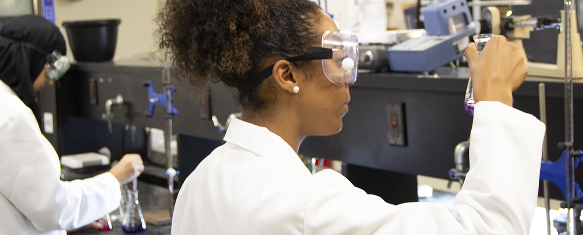 two students in lab coats and googles doing chemistry