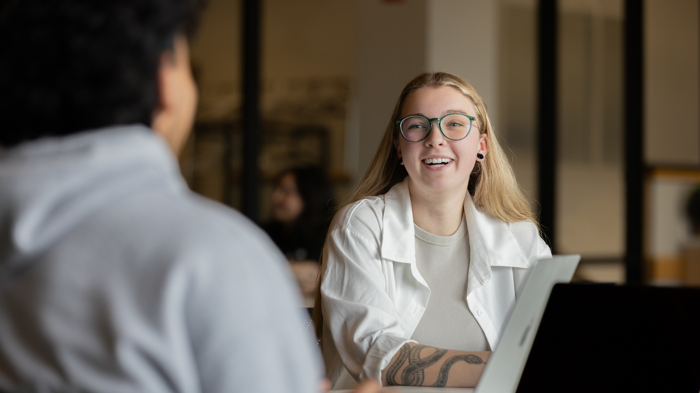 A smiling student wearing glasses talks with another person across a table, with an open laptop in front of her.