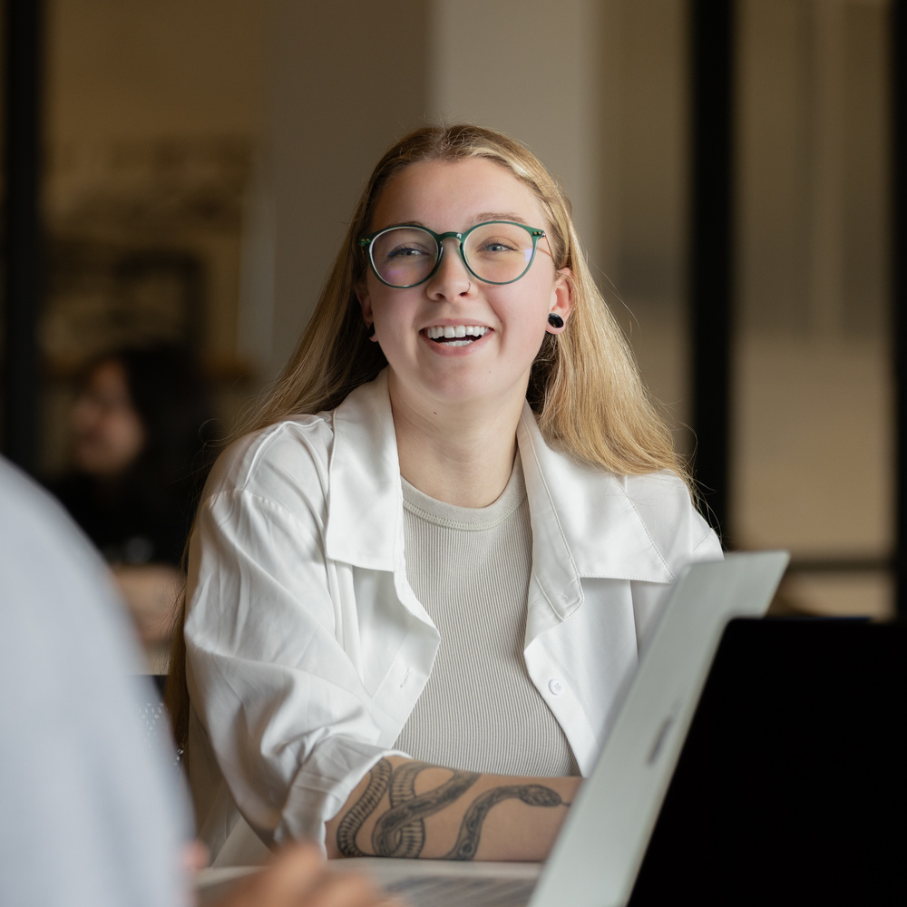 A smiling student wearing glasses talks with another person across a table, with an open laptop in front of her.