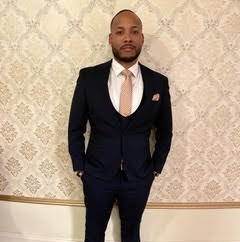 A gentleman in a dark suit and tie with his hands in his pockets in front of a wall with beige patterned wallpaper