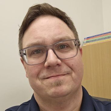 Headshot of John Pierce, a person with short brown hair and glasses, wearing a dark collared shirt and smiling in an indoor office space.