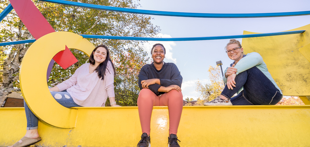 3 UMA students smiling and sitting on a campus sculptural installment on the Augusta campus