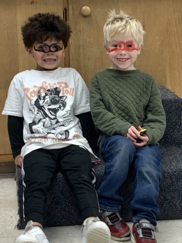 two boys sitting side by side. the one of the left is darker in coloring wearing upside down black glasses. the one on the right is blond with a green sweater wearing upside down red glasses