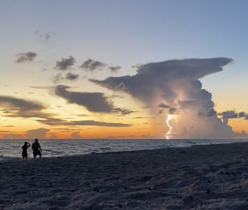 two people standing on a beach at sunset with a lightening bolt extending from the sky