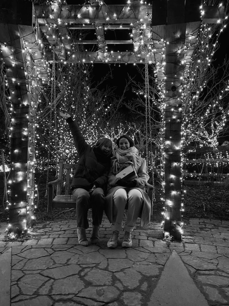 black and white photo of two girls sitting on a wooden swing surrounded by white lights