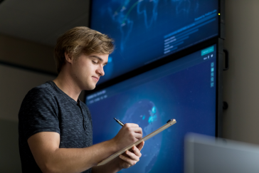 student writing on a clipboard in front of cybersecurity screen