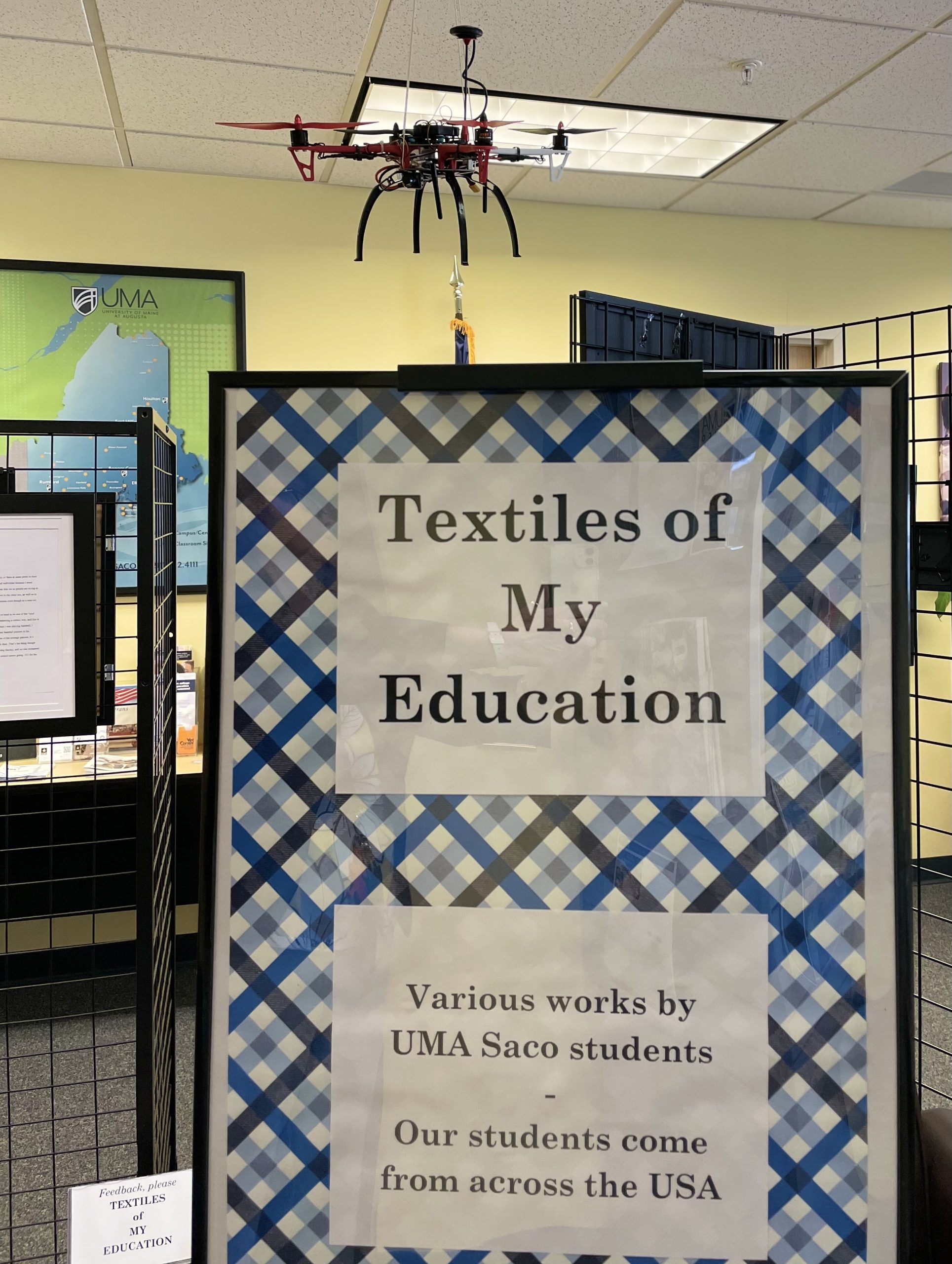 The entrance to the display of student work is a sign with a plaid background which reads "Textiles of my education. 