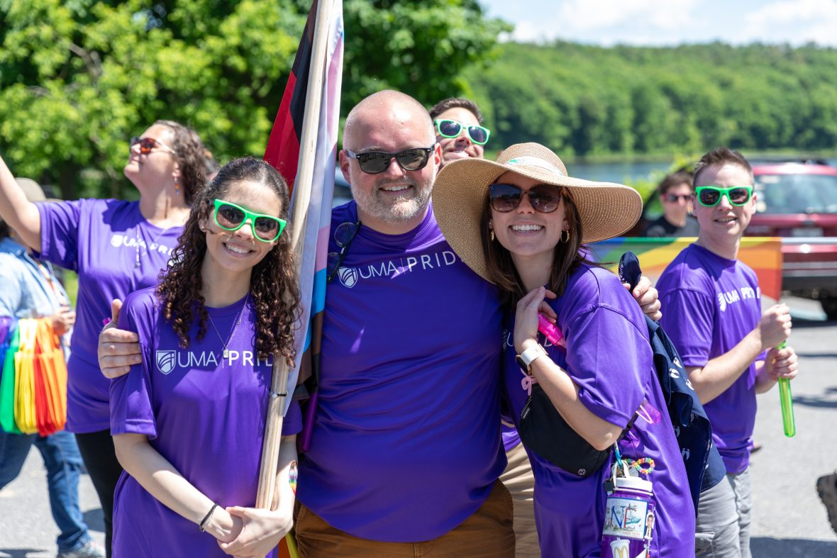 Three people wearing matching UMA Pride shirts and sunglasses pose for the camera. Other pride festivalgoers and trees are in the background.