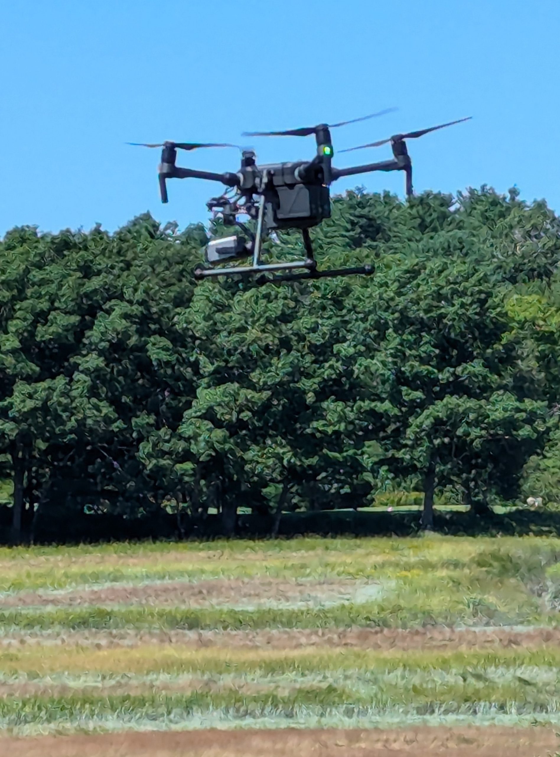 A drone hovers above a field with green trees in the background
