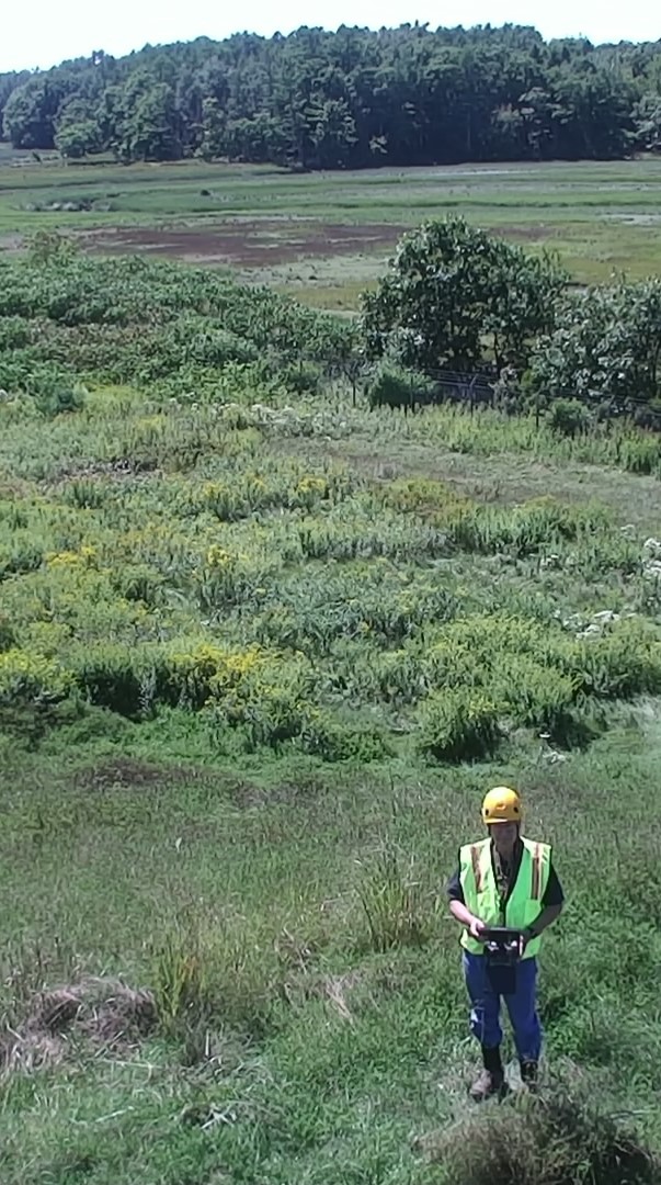 Dr. Daniel Leclair, wearing a hardhat and safety vest, stands in marshland using a drone’s ground control station