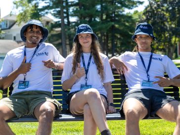 Three young people wearing blue hats, white t-shirts, and, ID badges sit on a park bench smiling and giving a thumbs up