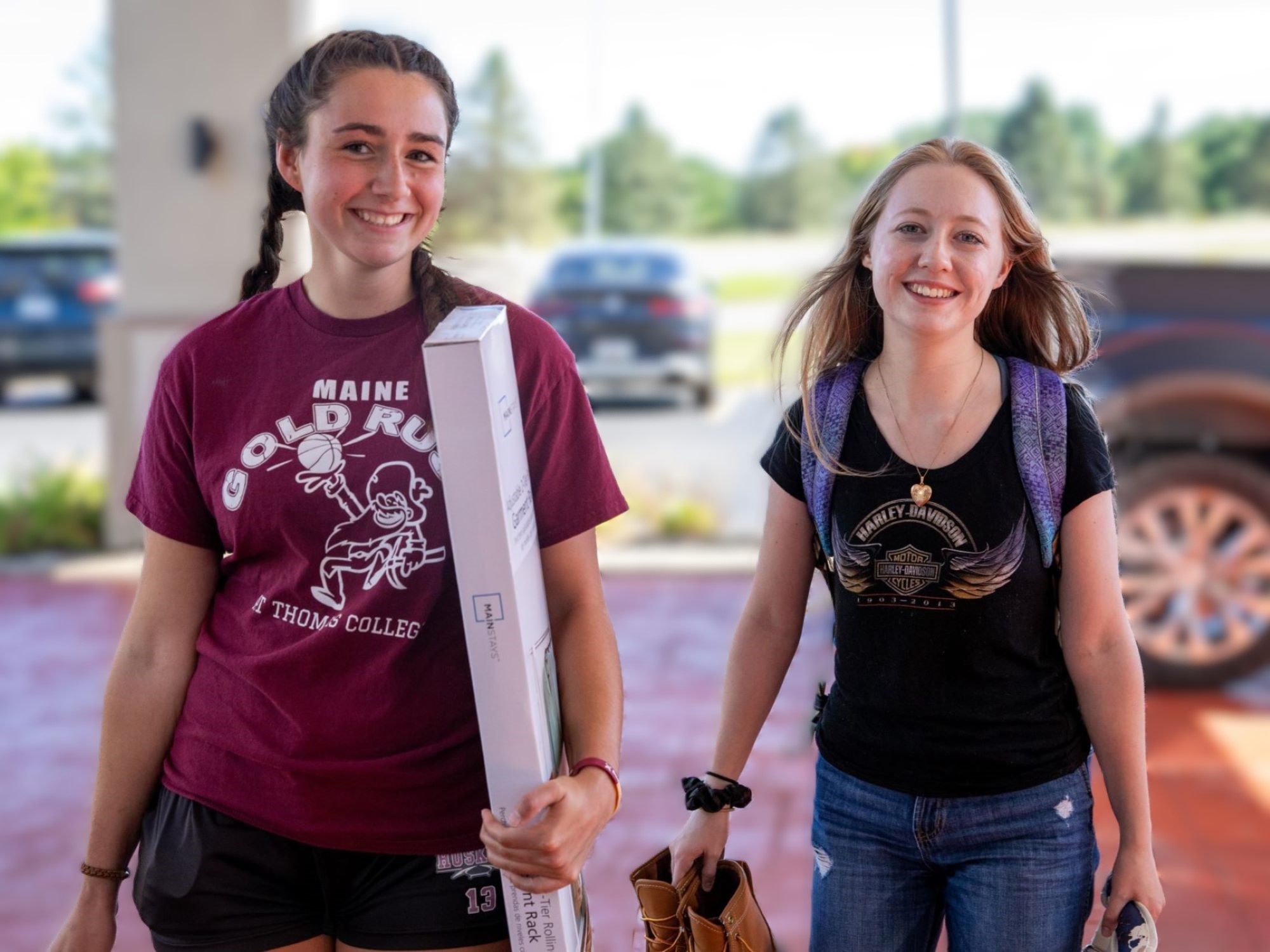 Two smiling young women carrying belongings as they move into student housing.
