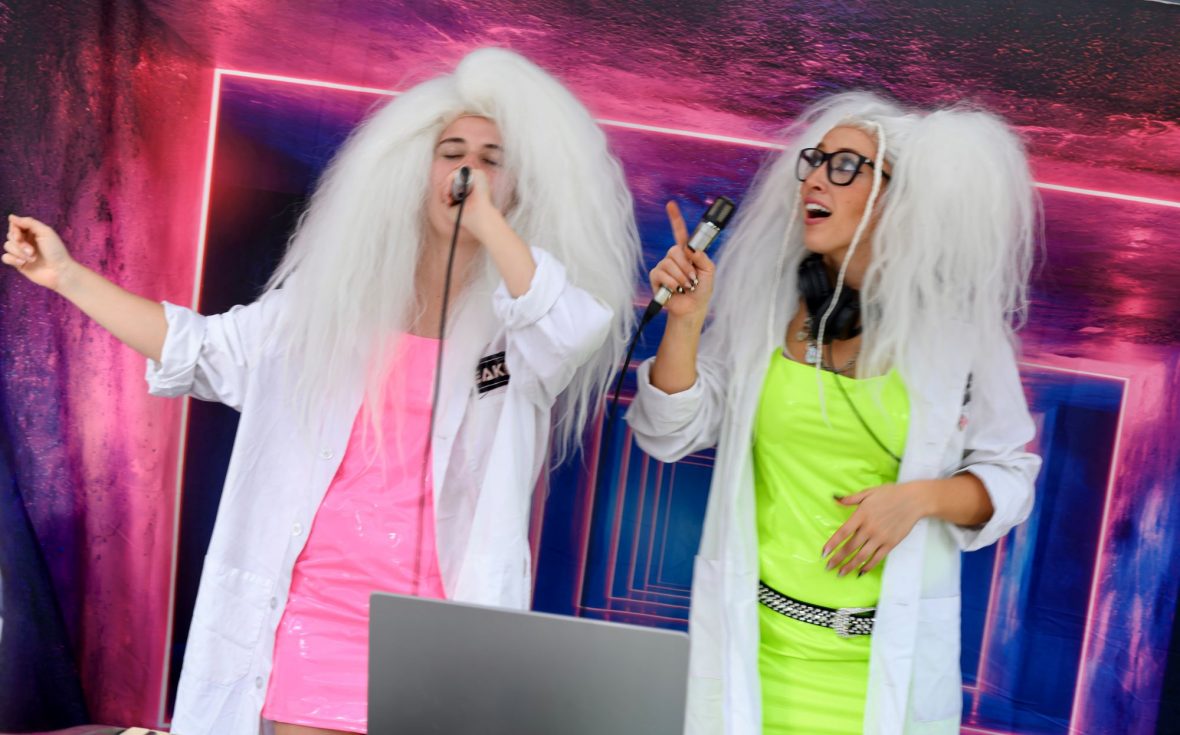 Two singers in large white wigs, neon dresses and white lab coats sing into microphones