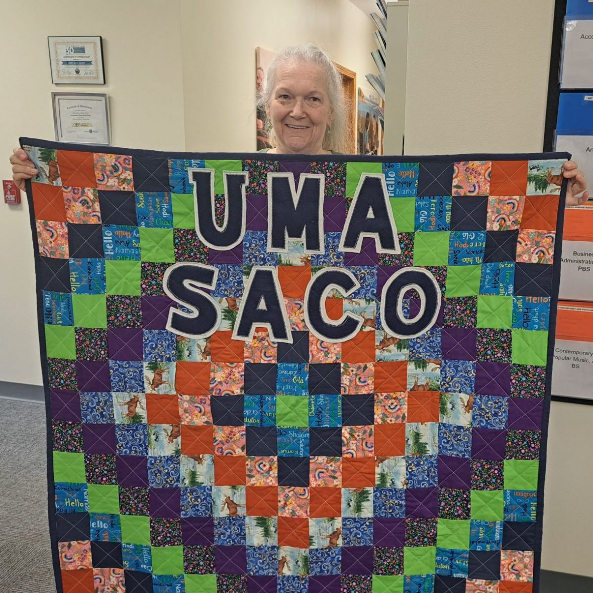 Brenda Plummer holds up a colorful handmade quilt inside the UMA Saco Center. The quilt features the words "UMA Saco" in large bold letters and includes bright fabric squares with Moose images, paisley patterns, flowers, and the word “Hello” in multiple languages. Brenda is smiling warmly, holding the quilt from behind.