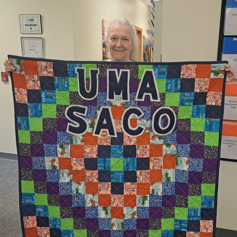 Brenda Plummer holds up a colorful handmade quilt inside the UMA Saco Center. The quilt features the words "UMA Saco" in large bold letters and includes bright fabric squares with Moose images, paisley patterns, flowers, and the word “Hello” in multiple languages. Brenda is smiling warmly, holding the quilt from behind.