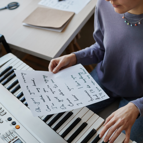 A person's hands hold a handwritten song lyric sheet over the keys of a white digital piano, with part of the piano visible at the bottom and a desk with papers and scissors in the blurred background.