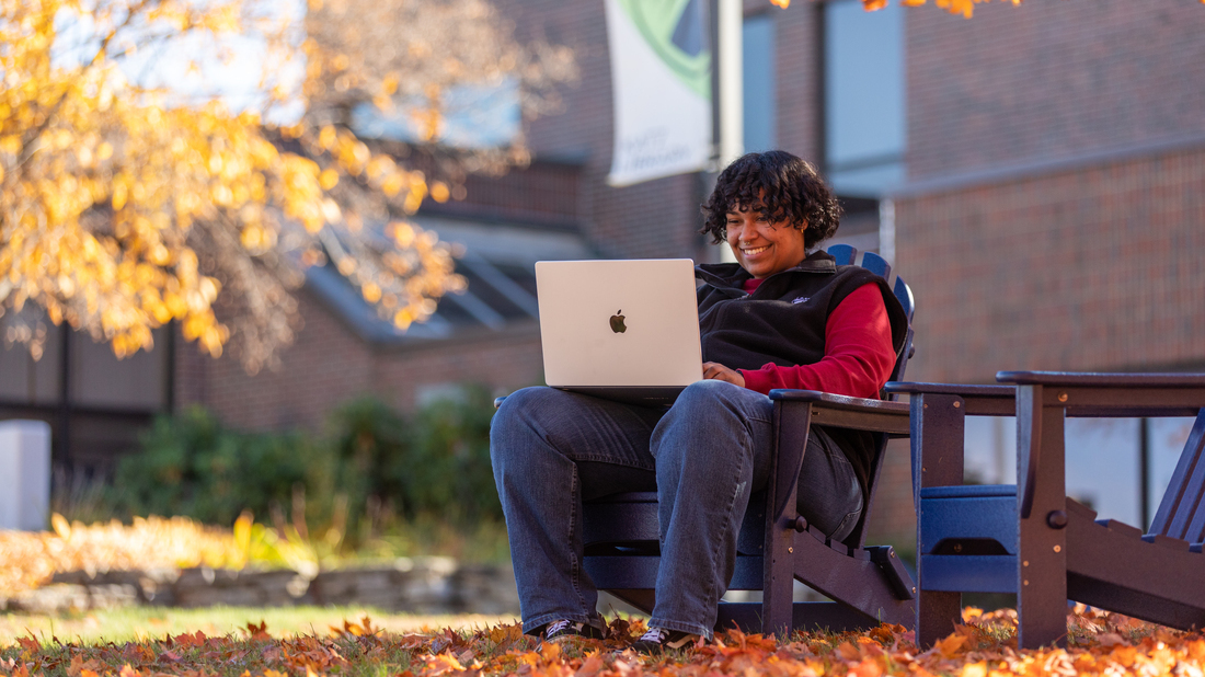 UMA student using laptop while surrounded by fall foliage on campus