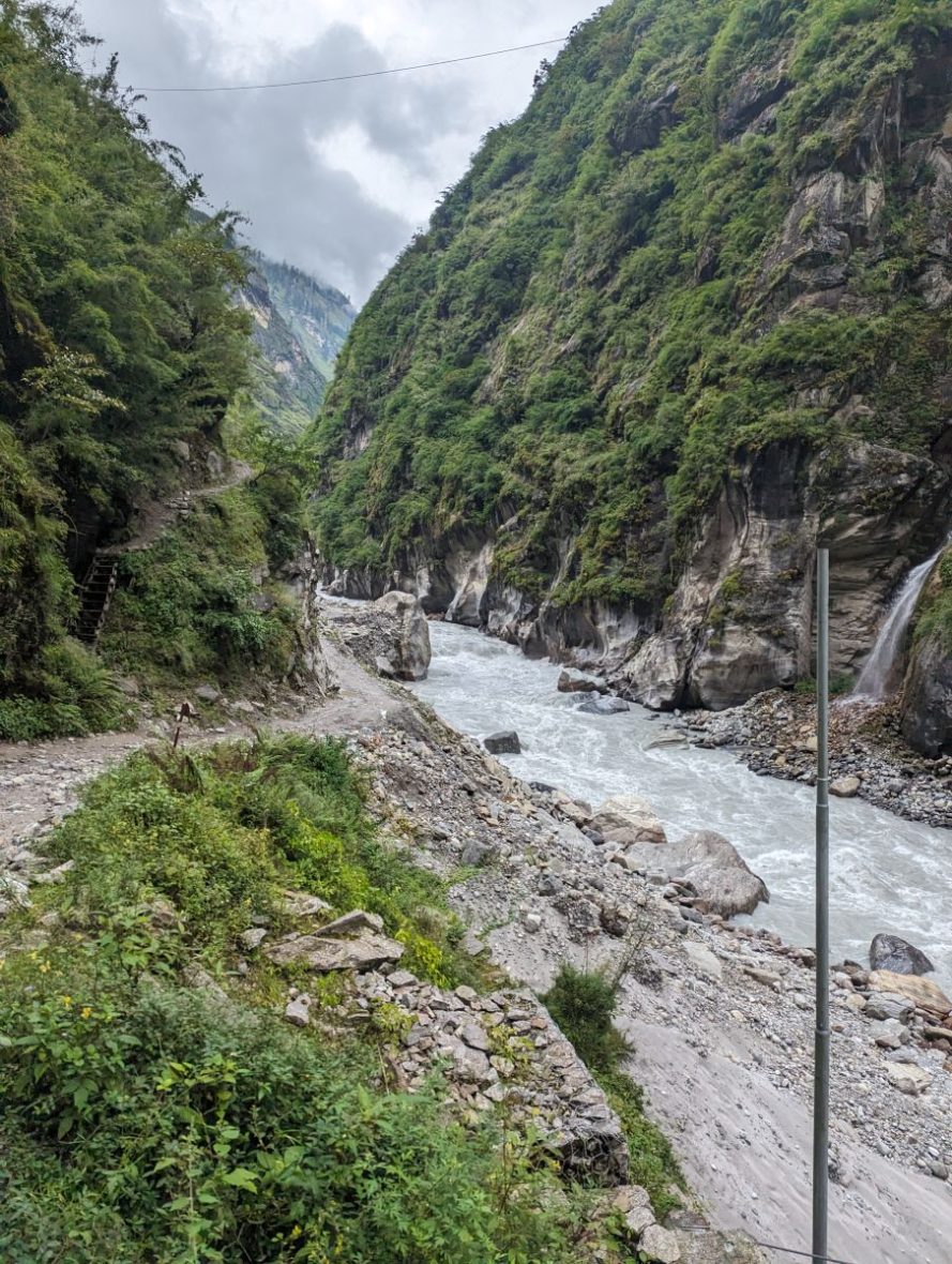 A narrow, rocky dirt road along side a rushing river in a narrow valley between two mountains