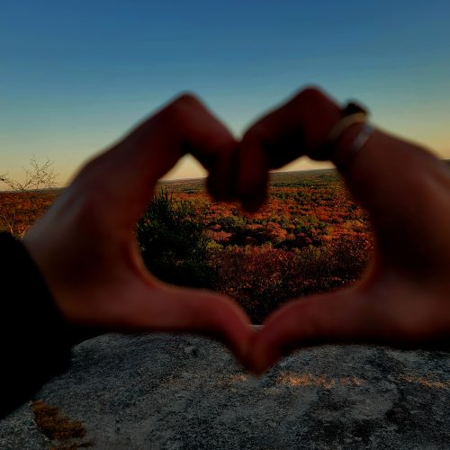 Framed by two hands forming a heart shape, a vibrant landscape unfolds at sunset, showcasing autumnal foliage in shades of orange, red, and green under a clear, blue sky.