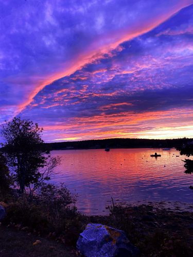 Vibrant sunset over a calm lakeshore with dramatic purple, pink, and orange clouds reflecting on the water,