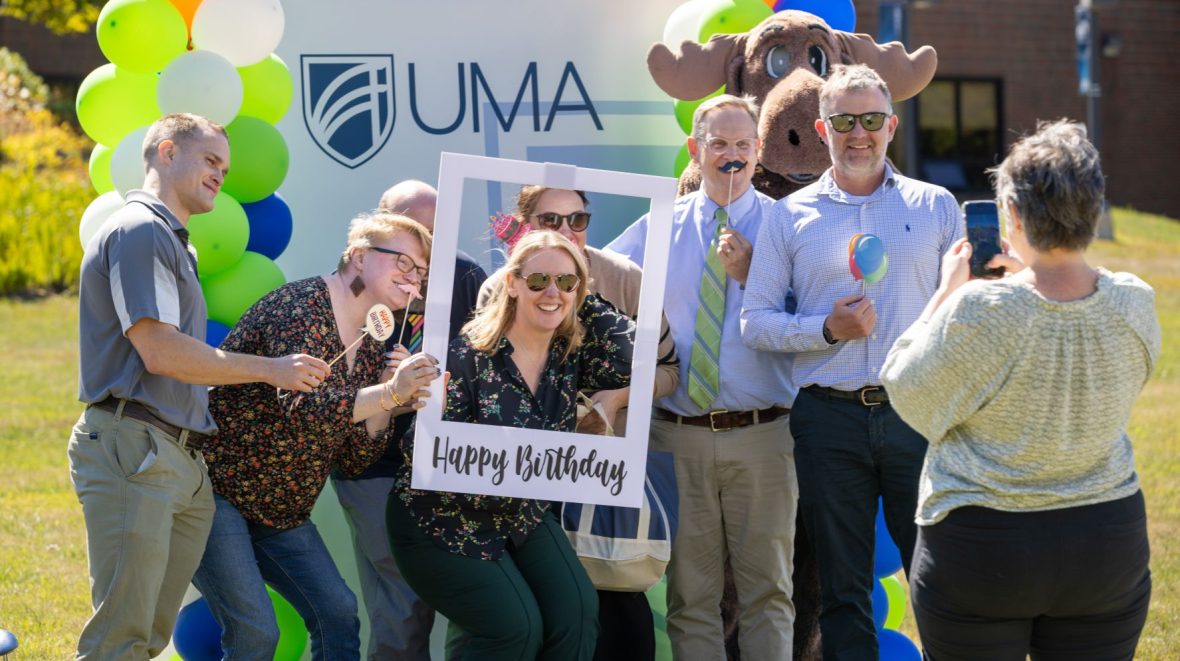 A woman takes a picture of a group of happy people holding props including a picture frame that says "Happy Birthday".
