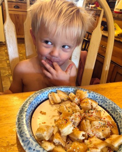 A young, shirtless boy with messy blond hair looks to his left while putting his finger in his mouth, in front of a paper plate filled with bite-sized pieces of pastry topped with sprinkles and syrup.