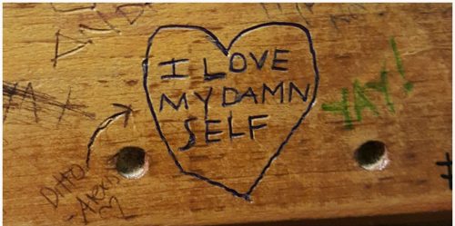 Close-up of a heart with the words "I LOVE MY DAMN SELF" written inside in dark blue marker on a weathered wooden surface, with other graffiti and two holes visible.