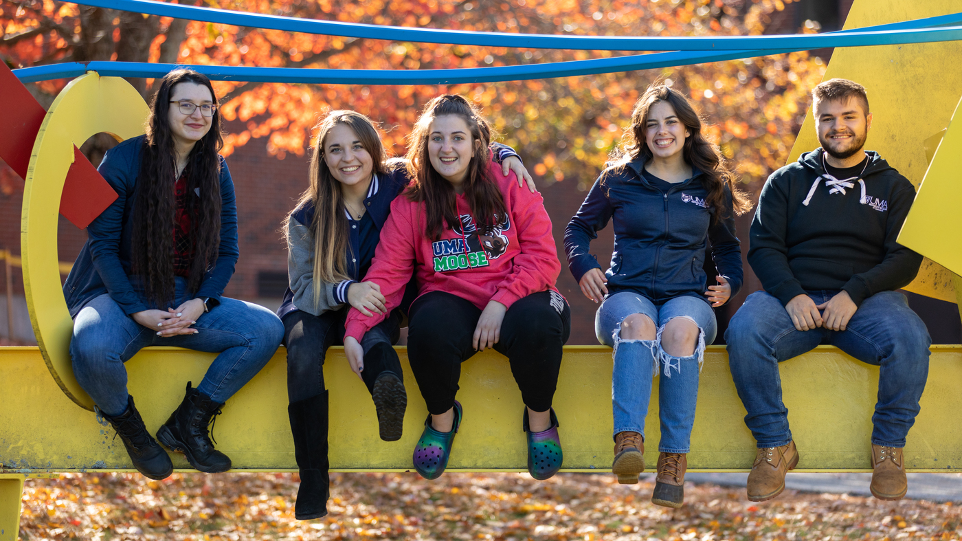 Five smiling UMA students sit on a yellow sculpture outdoors on a fall day with colorful leaves behind them.