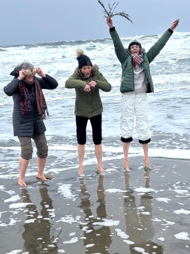 Three women stand at the edge of the ocean with foamy waves lapping at their feet. All three are wearing coats and pants, with two of them having removed their shoes, and the third wearing ballet flats.