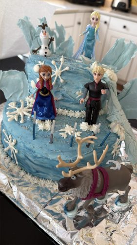 A two-tiered blue birthday cake decorated with icy textures, white frosting snowflakes, and miniature figures of characters from the movie Frozen, including Elsa, Anna, Kristoff, Olaf, and Sven the reindeer.