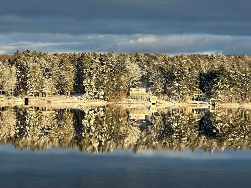 A snow-covered evergreen forest lines a calm lake, reflecting the trees and a few lakeside structures.