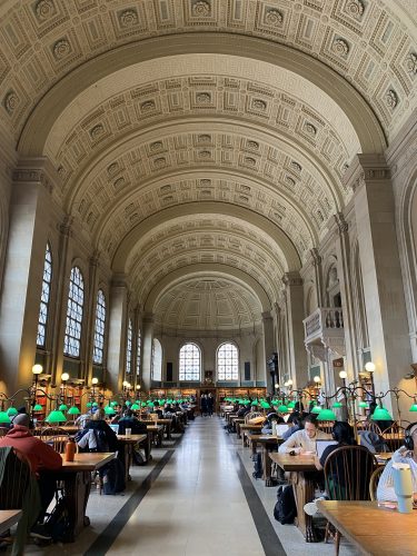 Rows of people sit at long wooden tables in a grand library hall with a highly ornate, domed ceiling and green desk lamps.