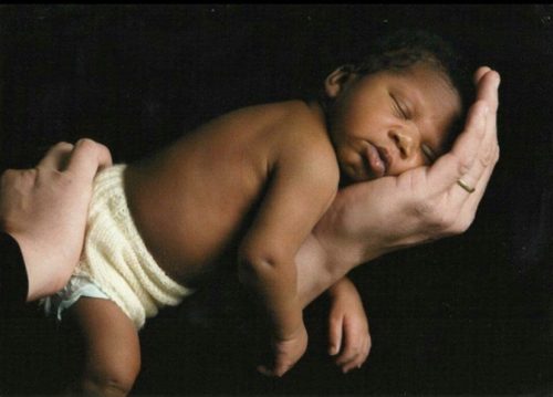 A sleeping infant with dark skin, wearing a knitted diaper cover, rests in the cradling palm of a hand against a black background.