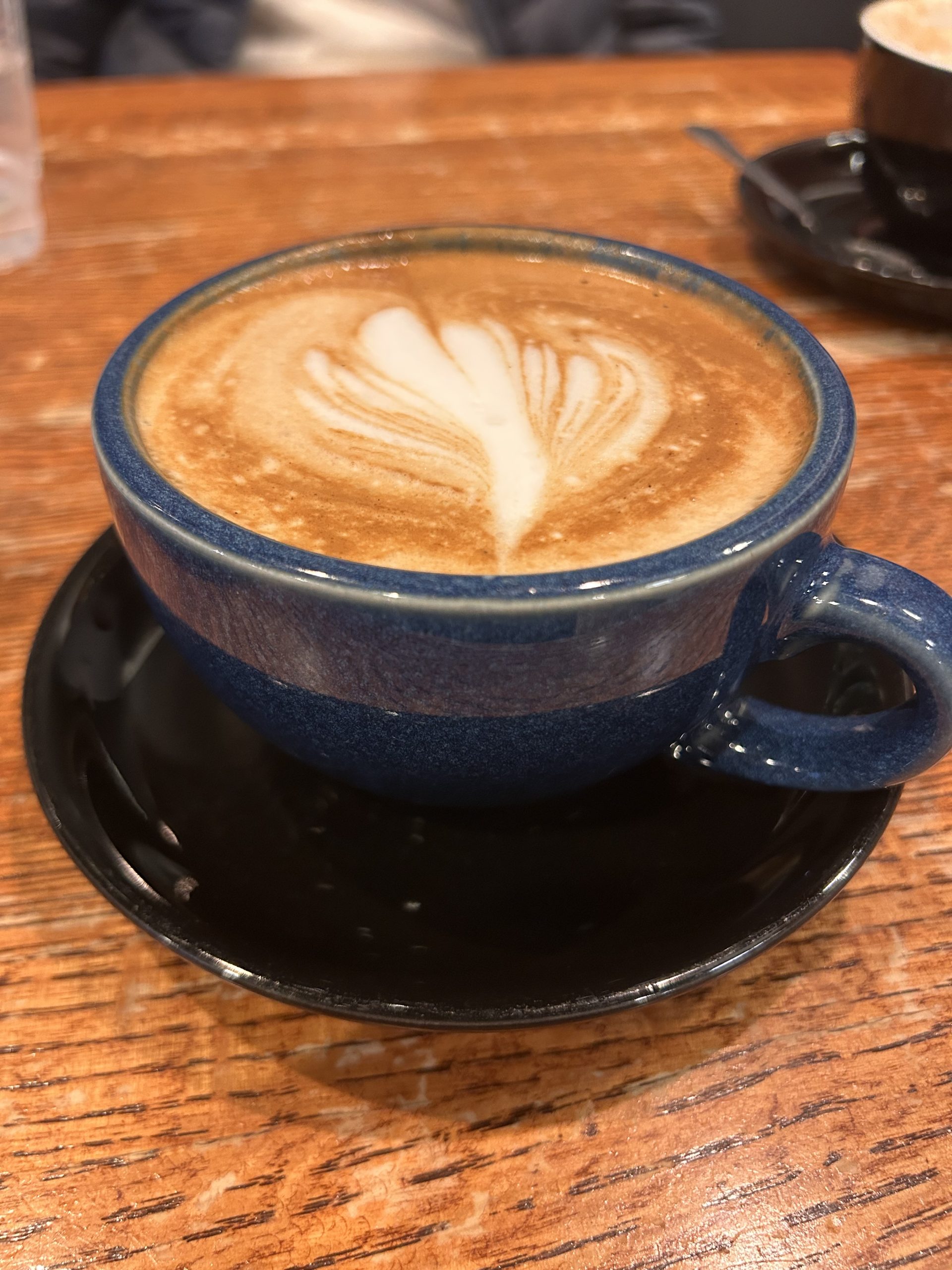 A close-up view of a blue mug filled with latte art sits on a black saucer atop a wooden table.
