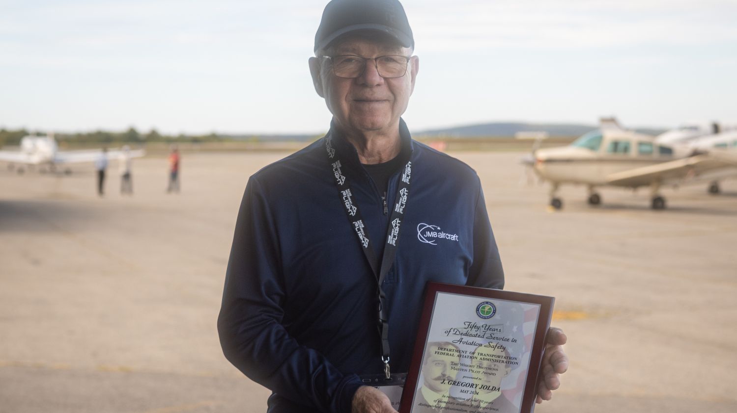 Pilot and UMA Professor Greg Jolda poses with his FAA Wright Brothers Master Pilot Award while standing on a tarmac with small planes in the background