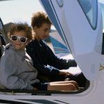 Two smiling children sit in the cockpit of a small airplane