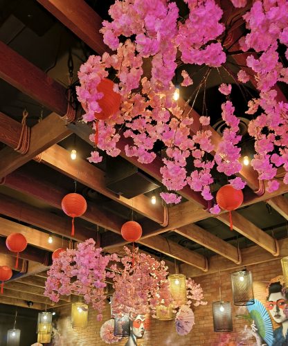 An interior view looking up at a ceiling decorated with pink artificial cherry blossoms, red paper lanterns, and exposed wooden beams, with industrial-style light fixtures hanging below and a wall featuring artwork of faces.