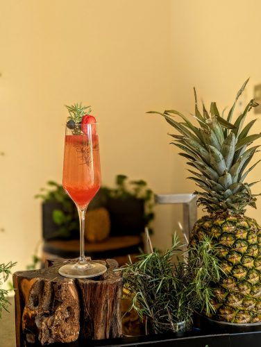 A tall flute glass filled with a pink beverage garnished with a strawberry slice, a blueberry, and a sprig of rosemary sits on a rustic wooden stand next to a whole pineapple and more rosemary sprigs.