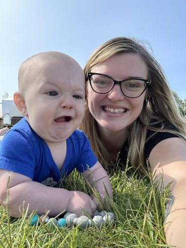 A baby wearing a blue shirt lies on the grass, making a funny face, next to a woman wearing glasses, who is smiling at the camera.