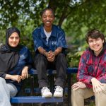 Three UMA students sit together at a blue picnic table outdoors on the Augusta campus. Two students wear UMA Ambassador shirts, and all are smiling under the shade of green trees.