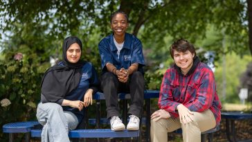 Three UMA students sit together at a blue picnic table outdoors on the Augusta campus. Two students wear UMA Ambassador shirts, and all are smiling under the shade of green trees.